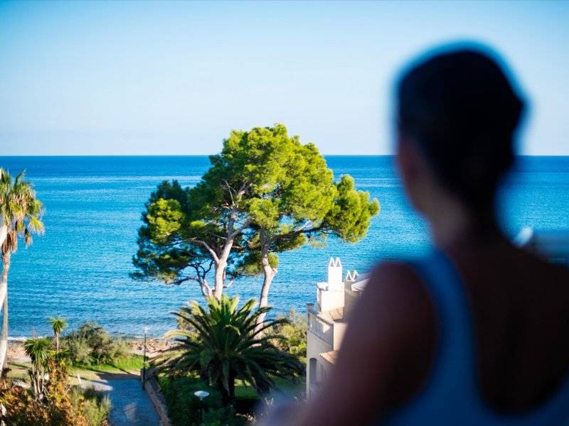 Frau blickt auf das Meer und einen Baum in der Ferne vom Balkon aus.