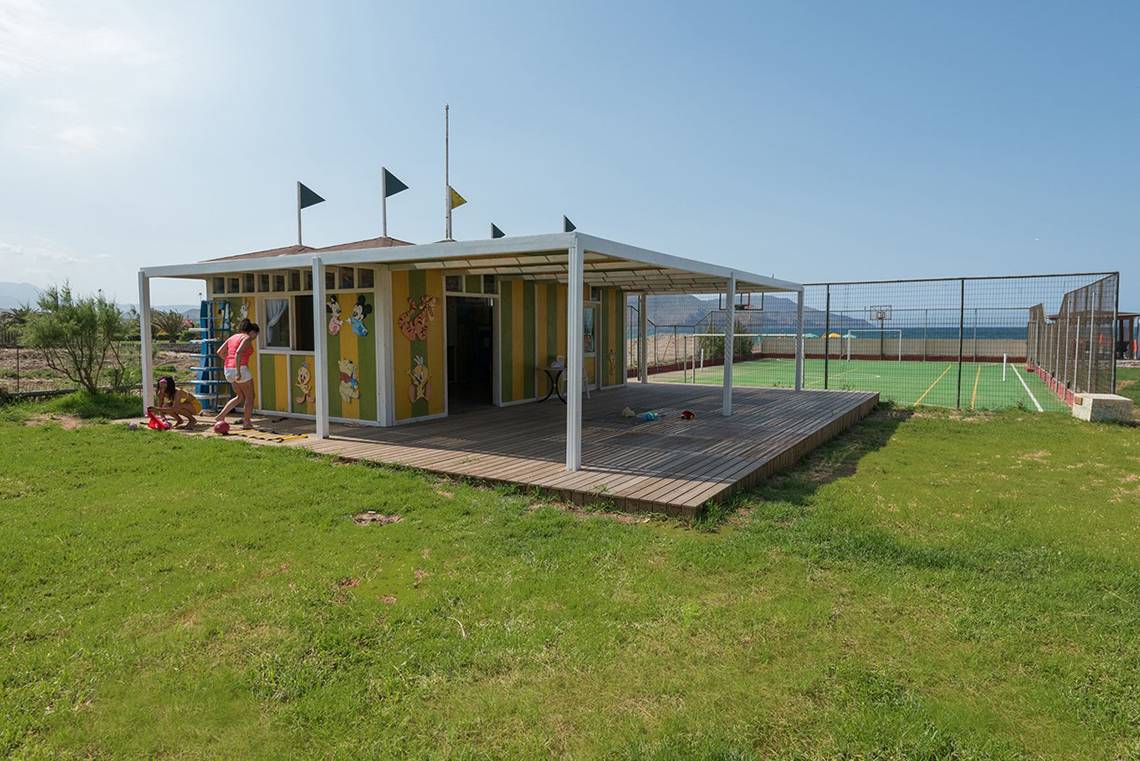 Außenansicht des Eliros Mare Hotel mit einem bunten Kinderspielhaus auf einem Sportplatz.