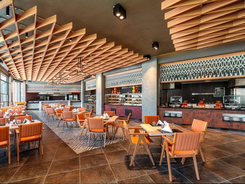 Innenbereich des Restaurants im Kirman Leodikya Resort mit modernen Holzmöbeln und einer offenen Küche.