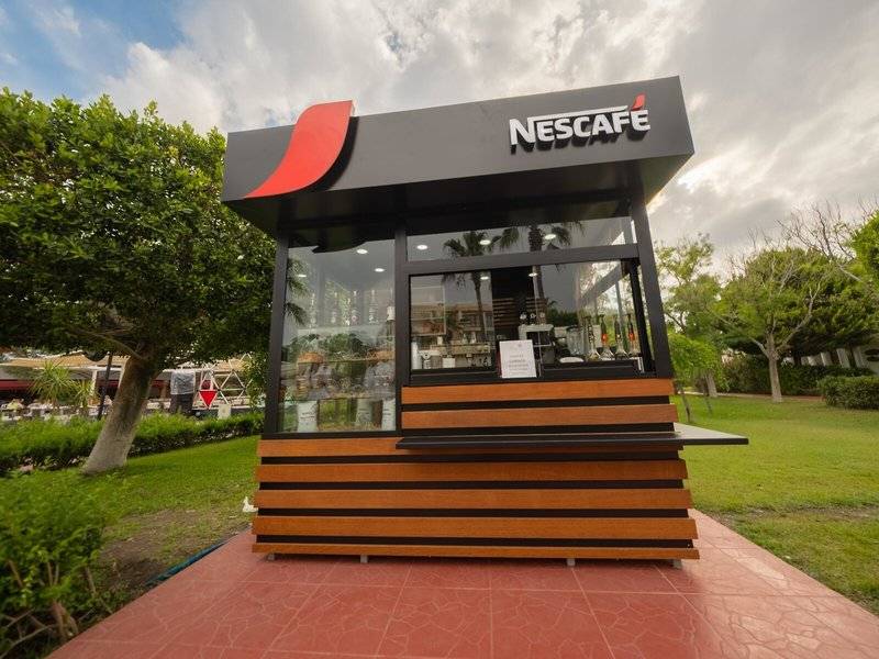 Außenansicht des Nescafé-Kiosks im Freien, umgeben von Bäumen und Rasen.