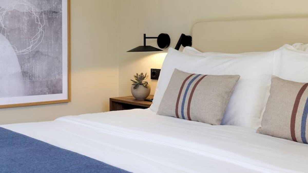 Bett im Domes Aulus Zante Hotel mit weißen Laken und Kissen, neben einem Nachttisch mit Pflanze.