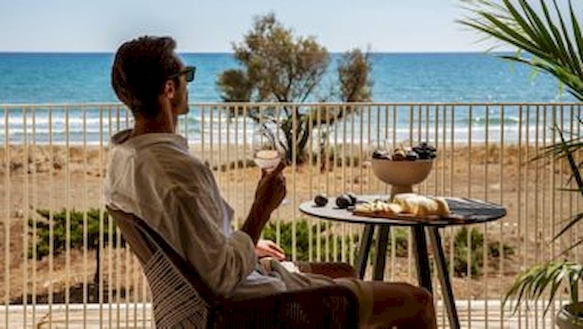 Ein Mann sitzt auf einem Balkon mit Blick auf das Meer und hält ein Glas Wein.