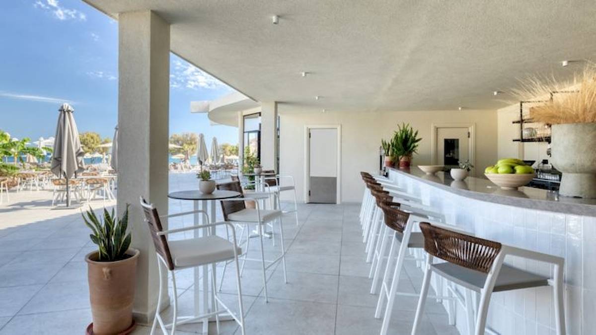 Innenansicht der Bar im Domes Aulus Zante Hotel mit weißen Möbeln, Tischen und Stühlen, Pflanzen und einer offenen Aussicht auf den Außenbereich.