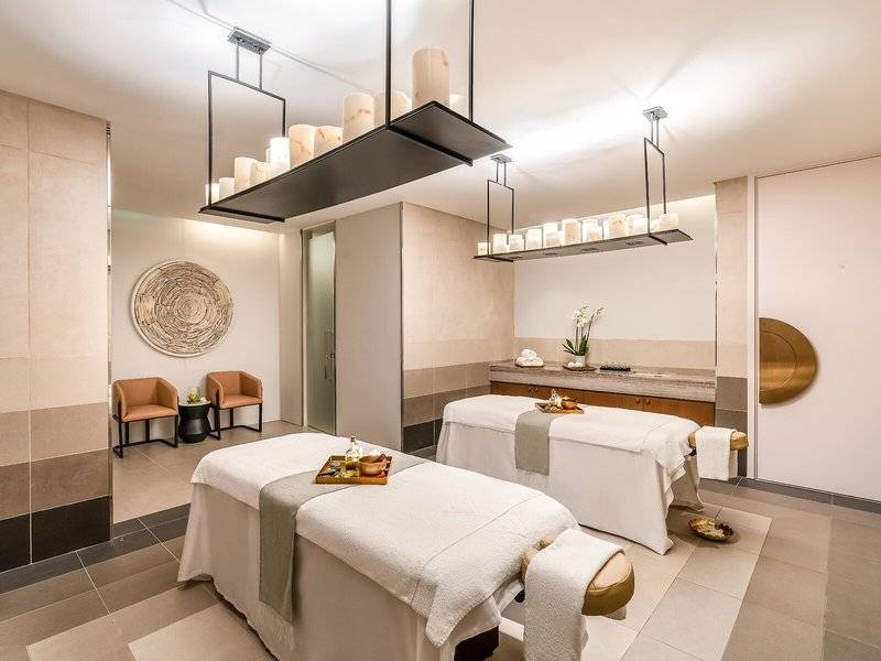 Behandlungsraum im Spa des Rixos Gulf Hotel Doha mit zwei Massagetischen, Sesseln und einem Wandbild.