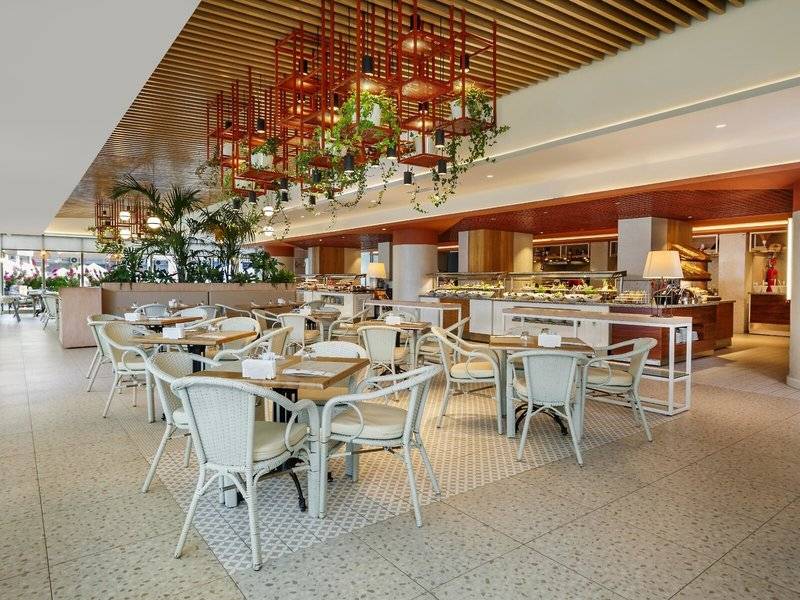 Innenbereich des Grand Park Lara Hotels mit geometrischen Mustern, Tischen und Stühlen, hängenden Pflanzen und einem Büffet.