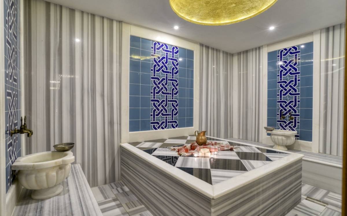Innenansicht des Hamams im Grand Park Lara Hotel mit blauen und weißen Fliesen, Marmorbadewanne und zwei Waschbecken.