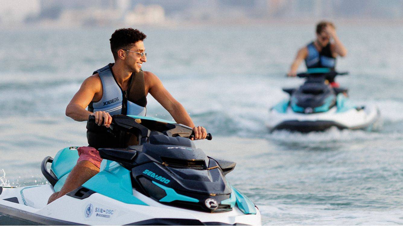 Zwei Personen auf Jetski im Wasser, eine vordere Person lächelt in die Kamera.