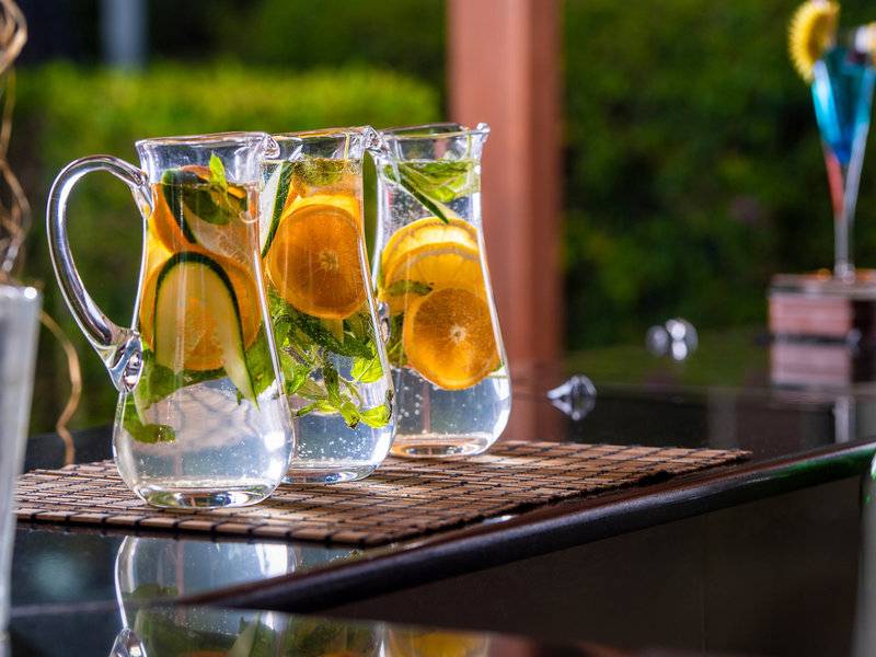 Drei Glaskruecke mit Wasser, Orangenscheiben, Gurkenscheiben und Minze auf einem Tisch.