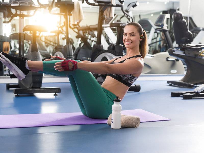 Frau im Fitnessstudio sitzt auf Matte und lächelt, während sie eine Beinübung macht