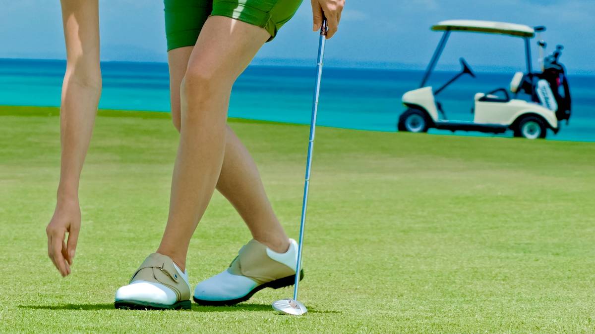 Golfspieler in grünen Shorts am Strand, ein Golfschläger haltend, mit einem Golfwagen im Hintergrund.