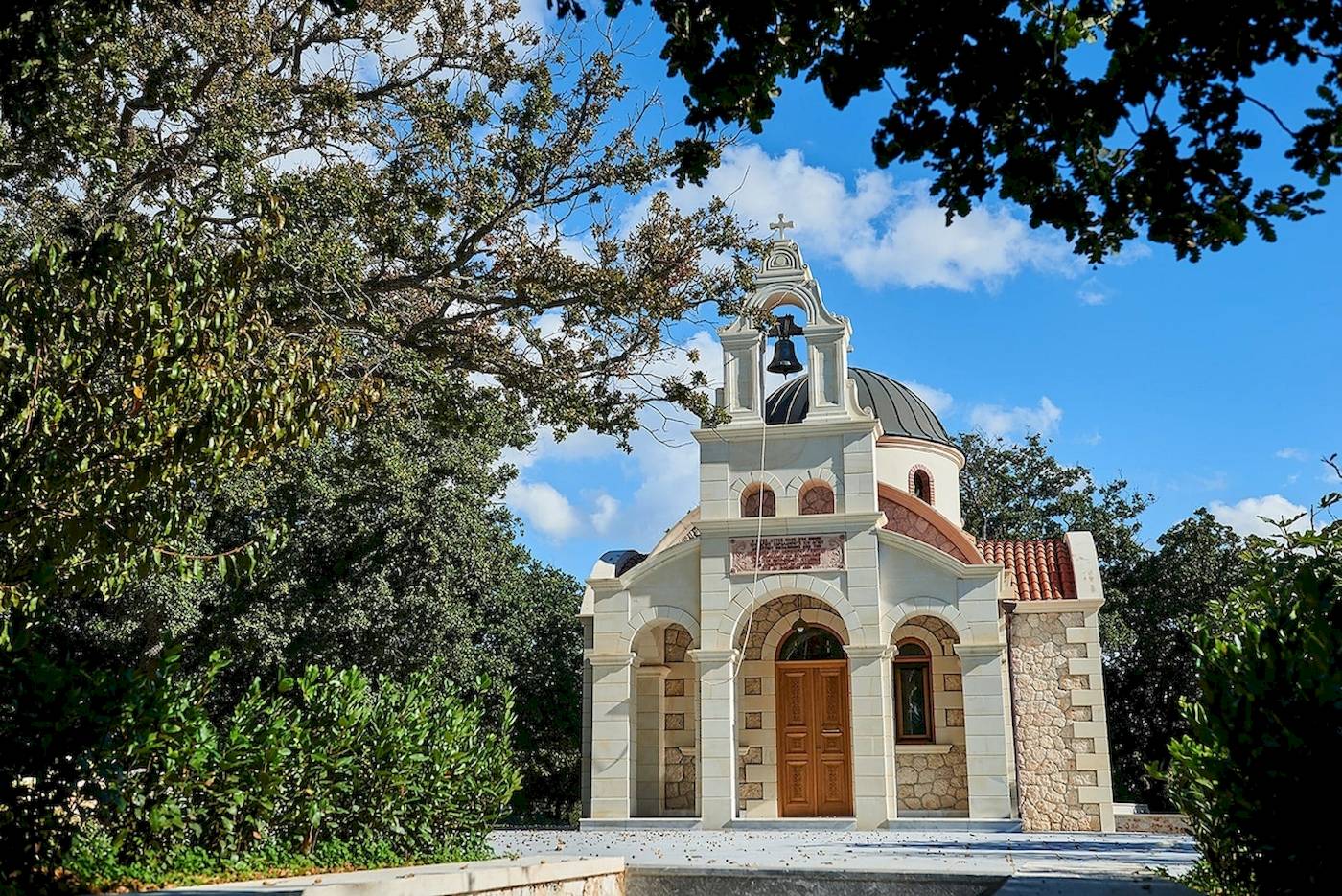 Kleine Kirche mit Glocke und Kuppel umgeben von Bäumen und Sträuchern