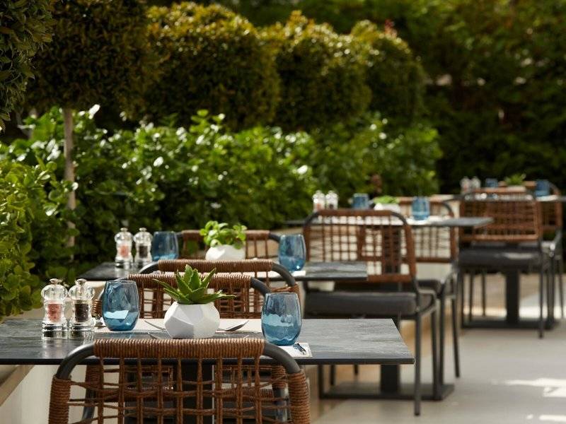 Außenbereich des Mare Paphos Hotels mit runden Tischen und Stühlen, blauen Gläsern und grünen Pflanzen.