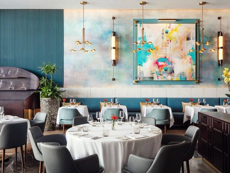 Innenraum des Restaurants im St. Regis Al Mouj Muscat Resort mit runden Tischen, modernen Möbeln und einer großen abstrakten Wandmalerei.