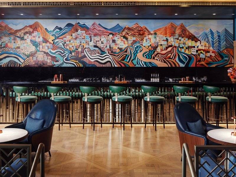 Innenansicht der Bar im St. Regis Al Mouj Muscat Resort mit buntem Wandgemälde und grünen Barhockern.