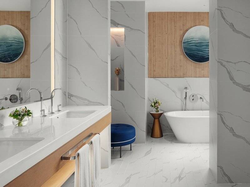 Badezimmer im St. Regis Al Mouj Muscat Resort mit Marmorwänden, zwei Waschbecken, Badewanne und Hocker.