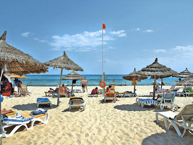 Strandszene mit vielen Sonnenschirmen und Liegestühlen, im Hintergrund das Hotel Paradis Palace.