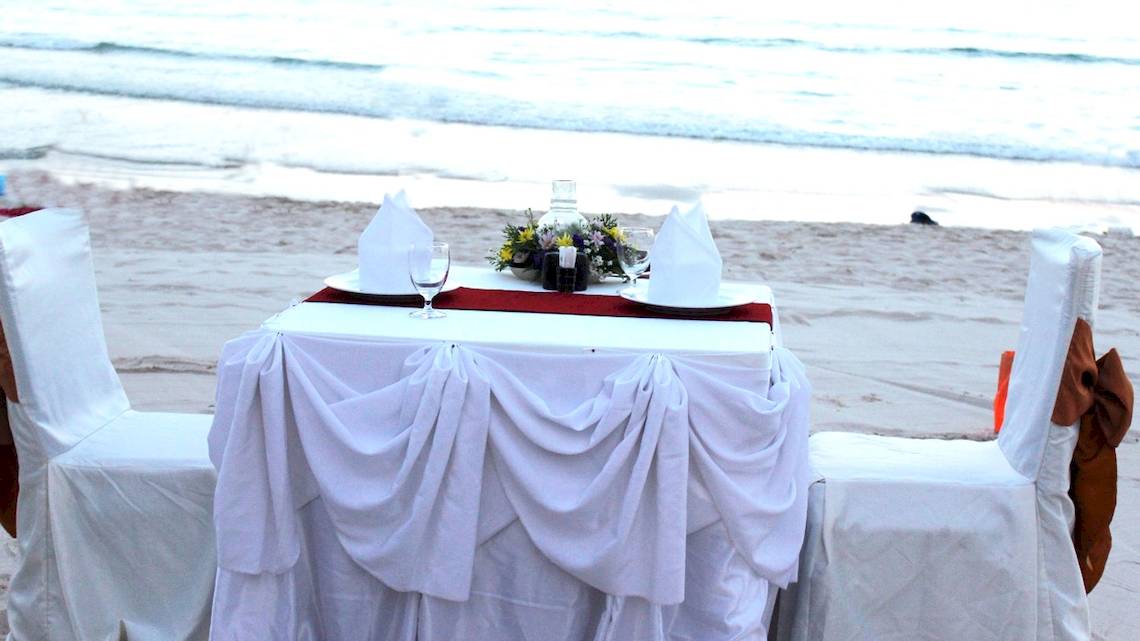 Ein Tisch auf dem Strand mit weißen Tischdecken und Blumen.