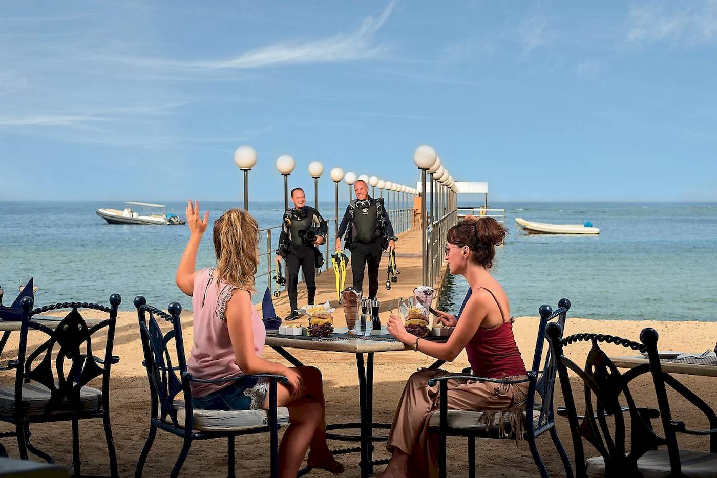 Scuba-Taucher, die auf einem Steg des Mövenpick Resort El Quseir stehen, während zwei Frauen am Tisch sitzen und essen.