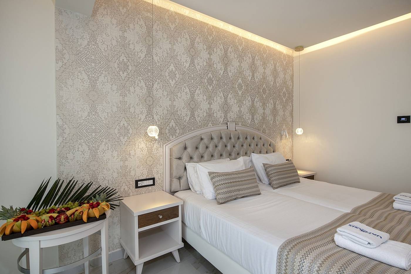 Schlafzimmer im Vantaris Luxury Beach Resort mit Bett, Nachttischen, Obstkorb und gemusterter Wand.