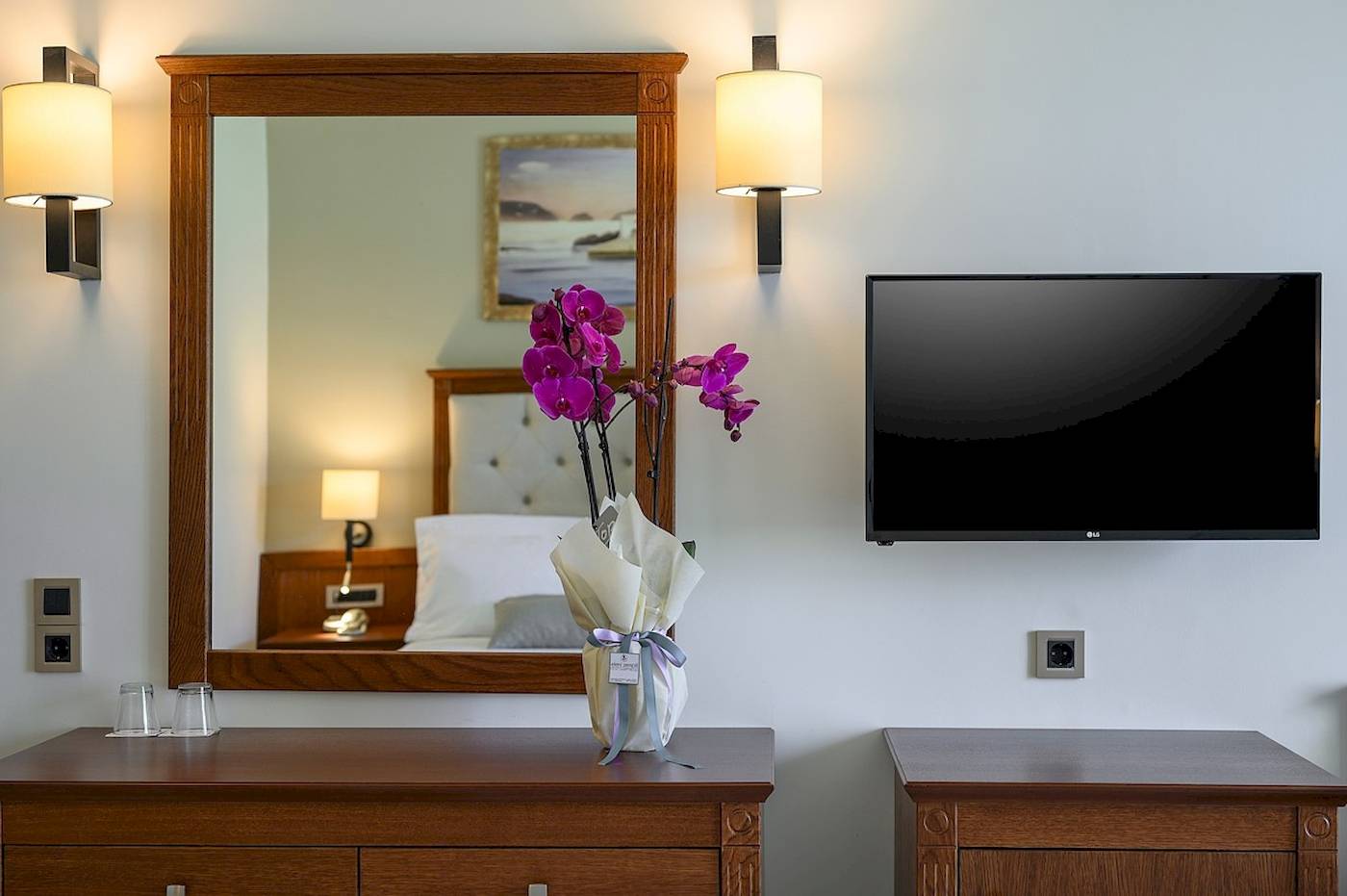 Ein Hotelzimmer mit einem Spiegel und einem Fernseher an der Wand, neben einem Nachttisch mit Blumen.