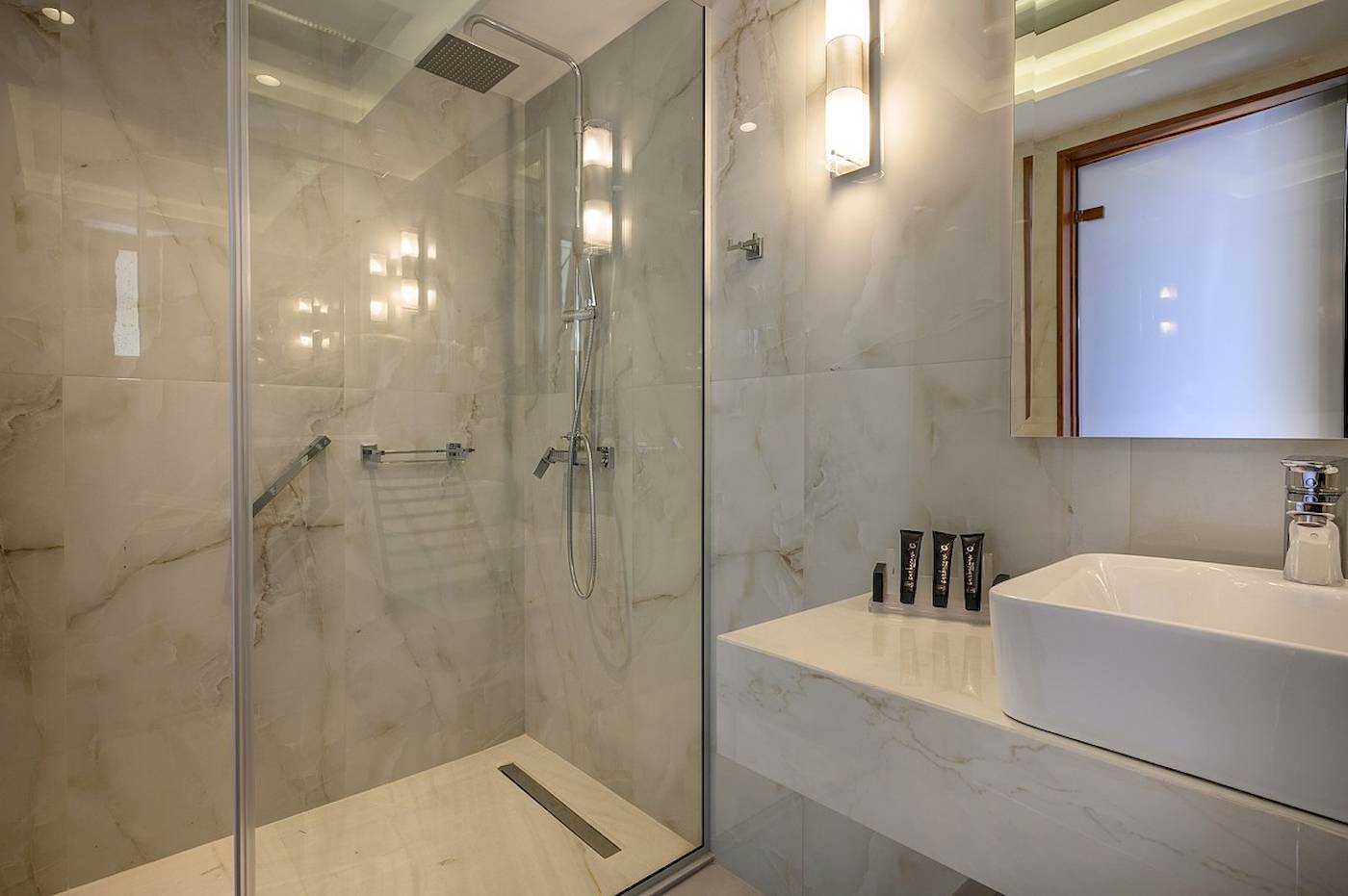 Ein elegantes Badezimmer im Vantaris Palace Hotel mit Marmorwänden, einer Glasdusche und einem Waschbecken.