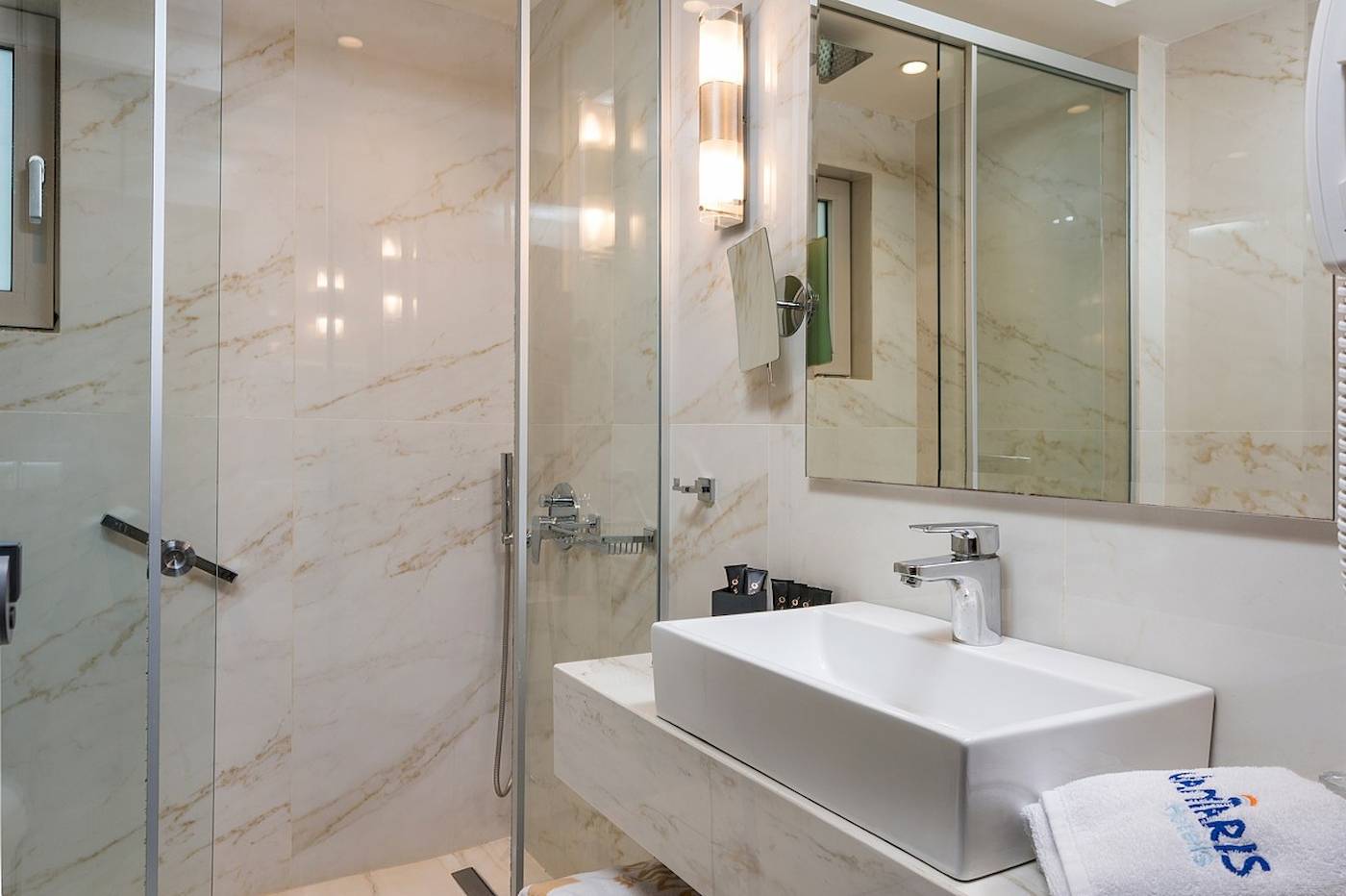 Badezimmer im Vantaris Palace Hotel mit Marmorwänden, Glasdusche und Waschbecken.