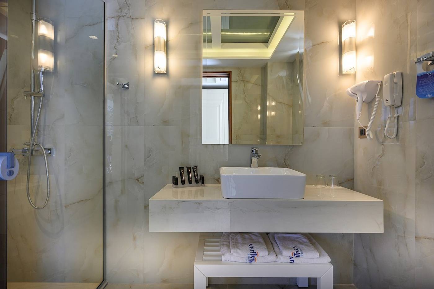Badezimmer im Vantaris Palace Hotel mit Marmorfliesen, Waschbecken, Spiegel und Föhn.