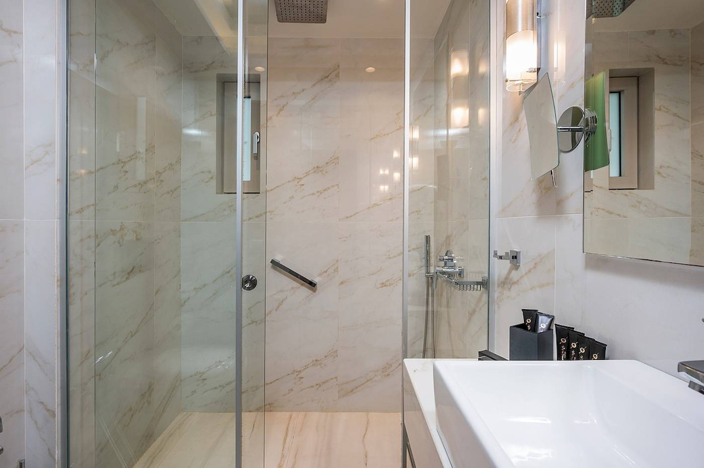 Badezimmer im Vantaris Palace Hotel mit Marmorwänden, Glasdusche und Waschbecken.