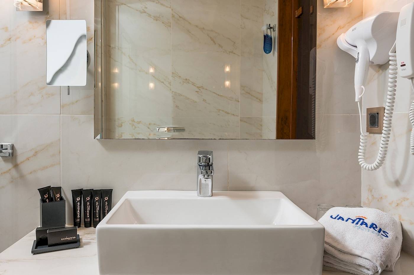 Badezimmer im Vantaris Palace Hotel mit Waschbecken, Spiegel und Handtuchtrockner.