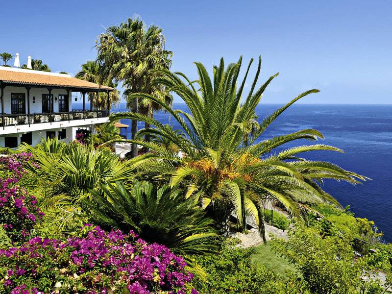 Außenansicht des Jardin Tecina Hotels mit Blick auf das Meer und eine Terrasse mit Palmen und Blumen.
