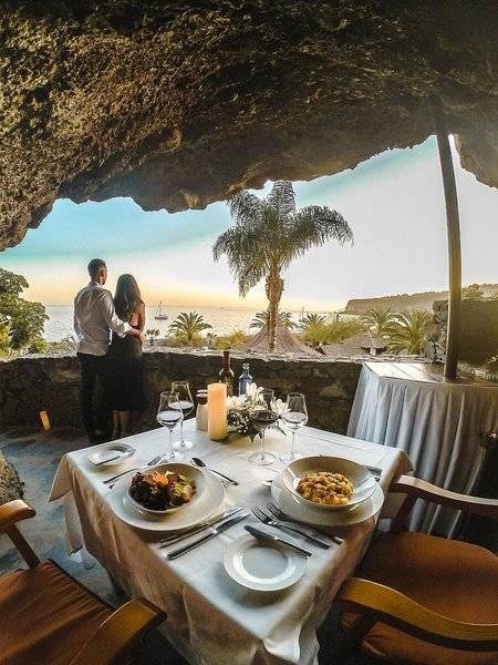 Ein Paar genießt ein Abendessen in einem Gartenrestaurant mit Meerblick, bei Sonnenuntergang.