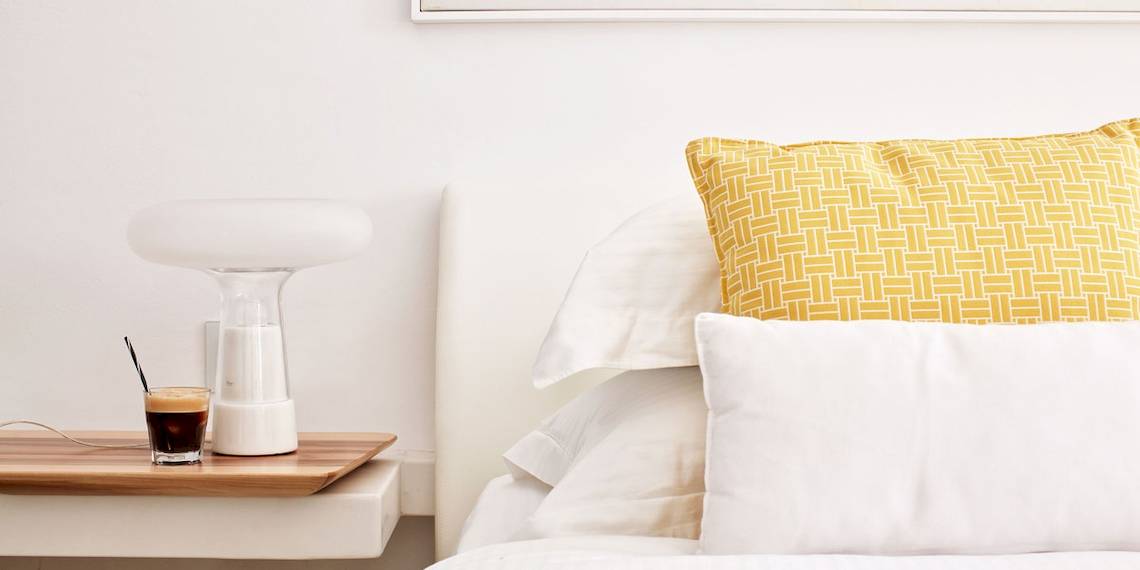 Hotelzimmer des Myconian Ambassador Relais & Chateaux mit Bett, gelbem Kissen und Nachttisch mit Lampe und Getränk.