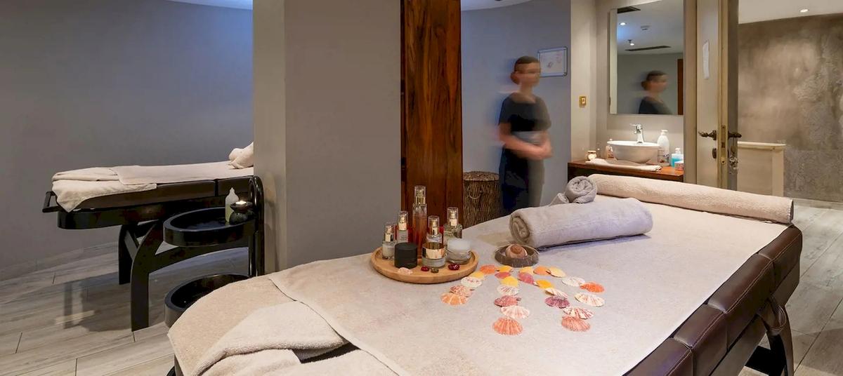 Massagebehandlungsraum im Cactus Village Hotel mit zwei Liegen, einer offenen Tür und einer Frau im Hintergrund.