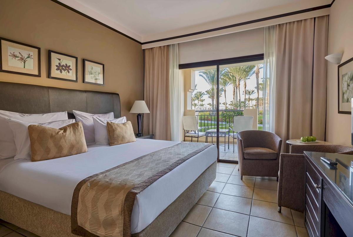 Schlafzimmer im Jaz Lamaya Resort mit einem großen Bett, Kissen und einem Balkon mit Blick auf Palmen.