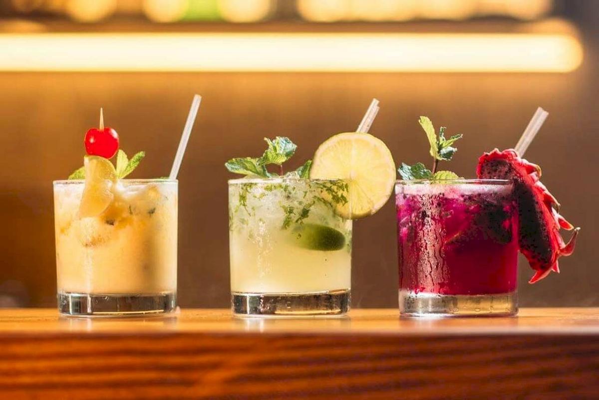 Drei Cocktails auf einem Holztisch, jeder mit verschiedenen Dekorationen wie Früchten und Kräutern.