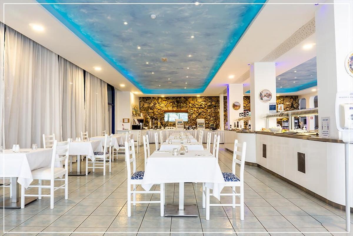 Restaurantbereich des Anonymes Strandhotel mit weißen Tischen und Stühlen, blauer Decke und Steinwand.