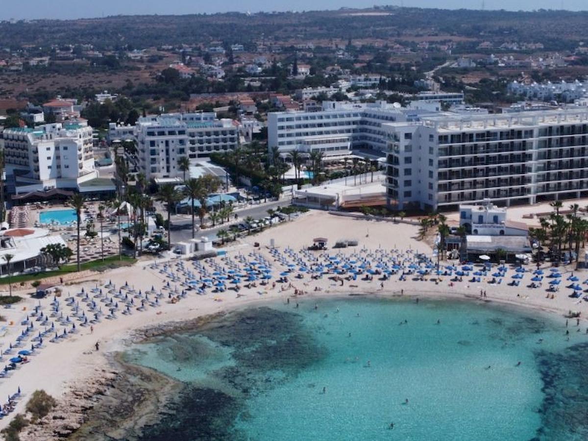 Luftaufnahme des Anonymous Beach Hotels mit vielen Gästen und blauen Sonnenschirmen am Strand.