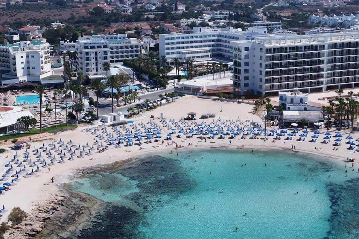 Luftaufnahme des Anonymous Beach Hotels mit vielen Gästen am Strand und blauen Sonnenschirmen.