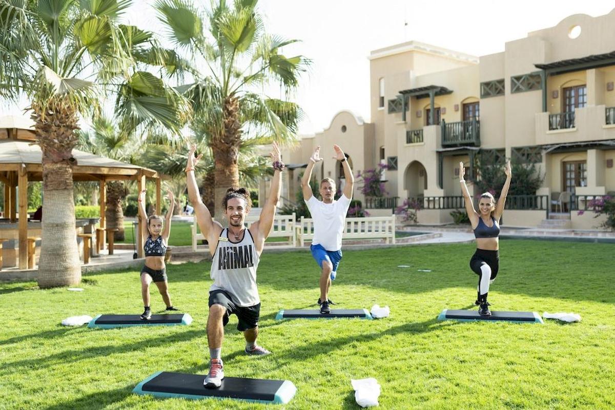 Fünf Personen machen Yoga im Freien auf dem Rasen im Freien vor einem Gebäude im Three Corners Rihana Resort.