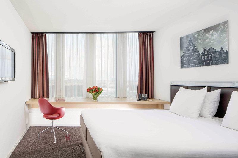 Innenansicht eines Zimmers im Casa Amsterdam Hotel mit einem Bett, Schreibtisch, rotem Stuhl und Fenstern.