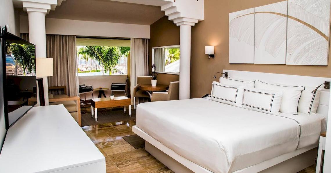 Innenansicht eines eleganten Zimmers im Melia Caribe Beach Resort mit einem großen Bett, Fernseher, Schreibtisch und Aussicht auf Palmen.