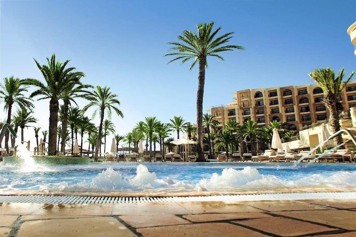 Außenpool des Mövenpick Resort & Marine Spa Sousse mit Palmen und Gebäude im Hintergrund.