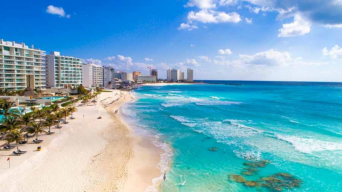 Luftaufnahme des Riu Cancun Hotels am Strand mit weißen Sand, blauem Ozean und Gebäuden im Hintergrund.
