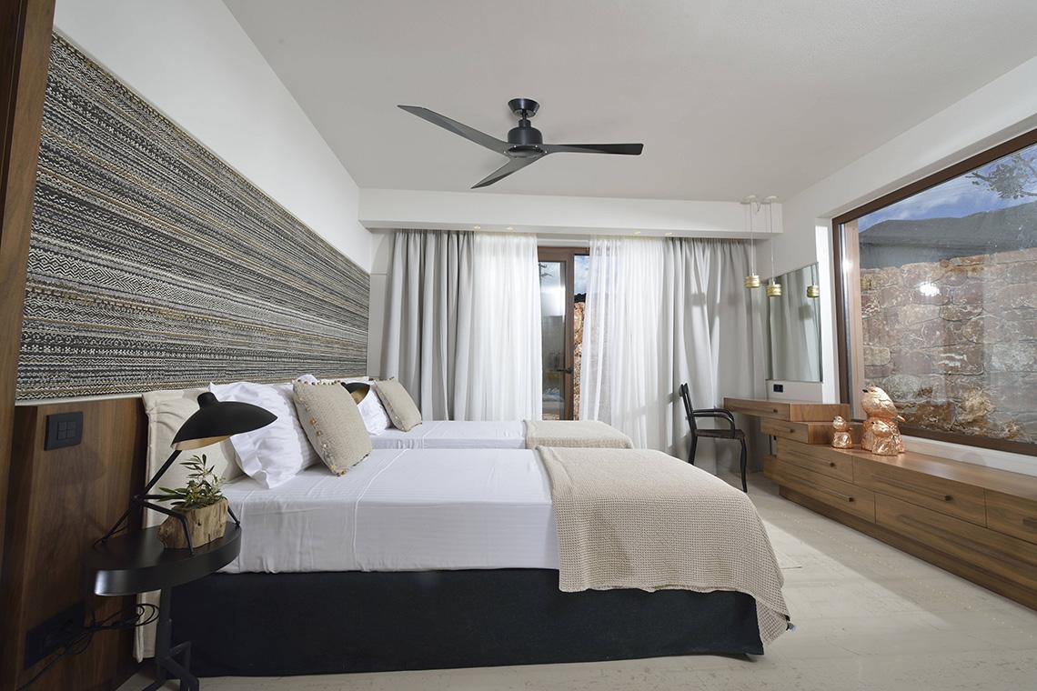 Schlafzimmer im Domes of Elounda, Autograph Collection mit zwei Betten, Deckenventilator und Steinmauer im Hintergrund.