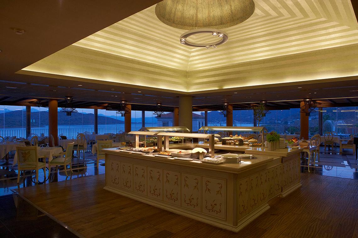 Innenansicht des Speisesaals im Domes of Elounda, Autograph Collection mit offenem Buffet, Tischen und Stühlen sowie großen Fenstern.
