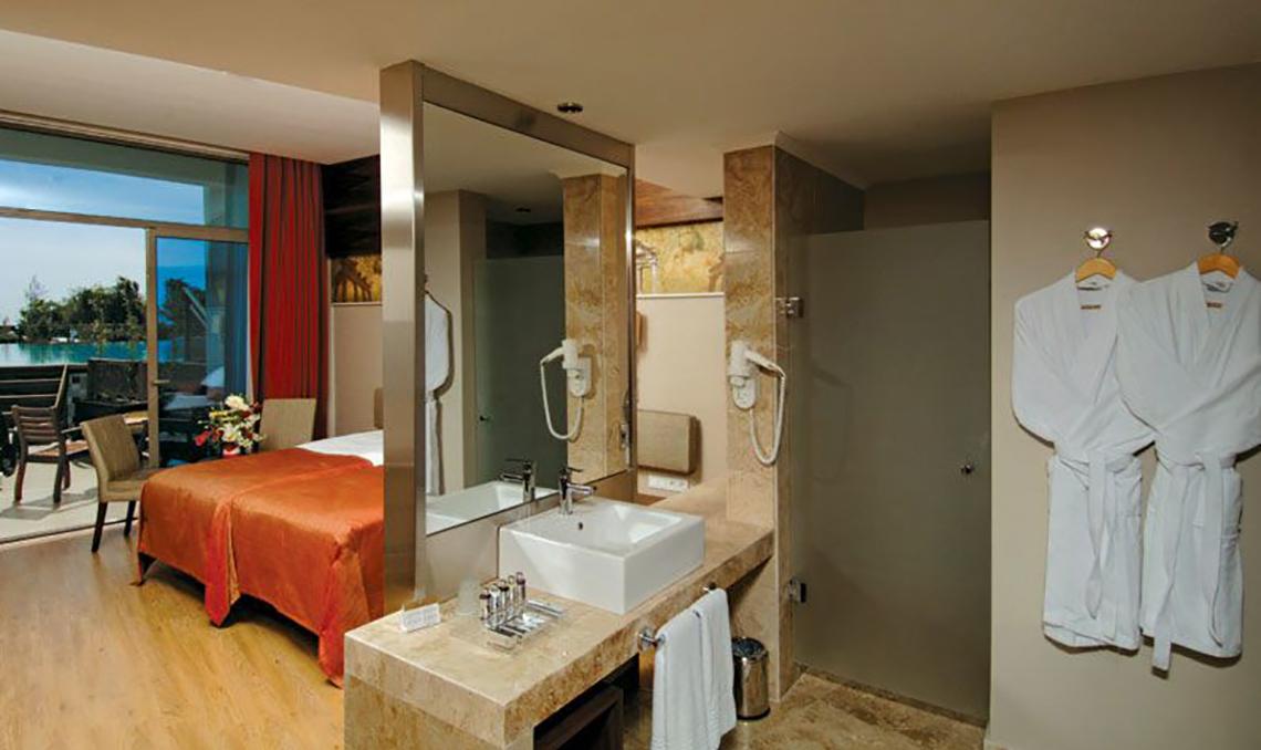 Badezimmer des Commodore Elite Suites & Spa mit Blick auf den Ozean, Bett, Spiegel, Waschbecken und Bademänteln.