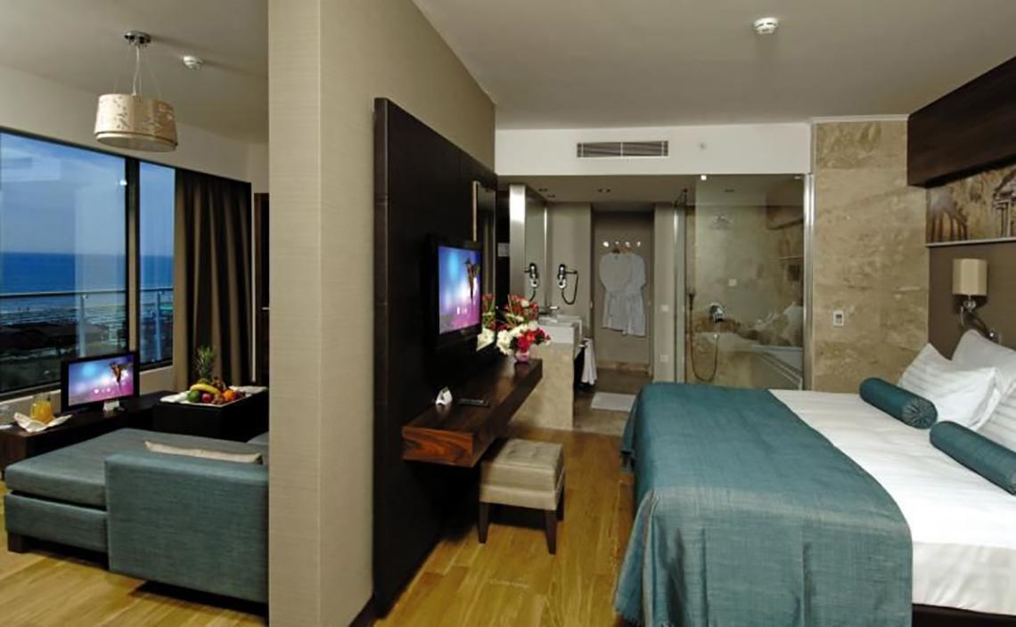Innenansicht eines Hotelzimmers im Commodore Elite Suites & Spa mit einem Bett, Fernseher, Tisch und Meerblick.