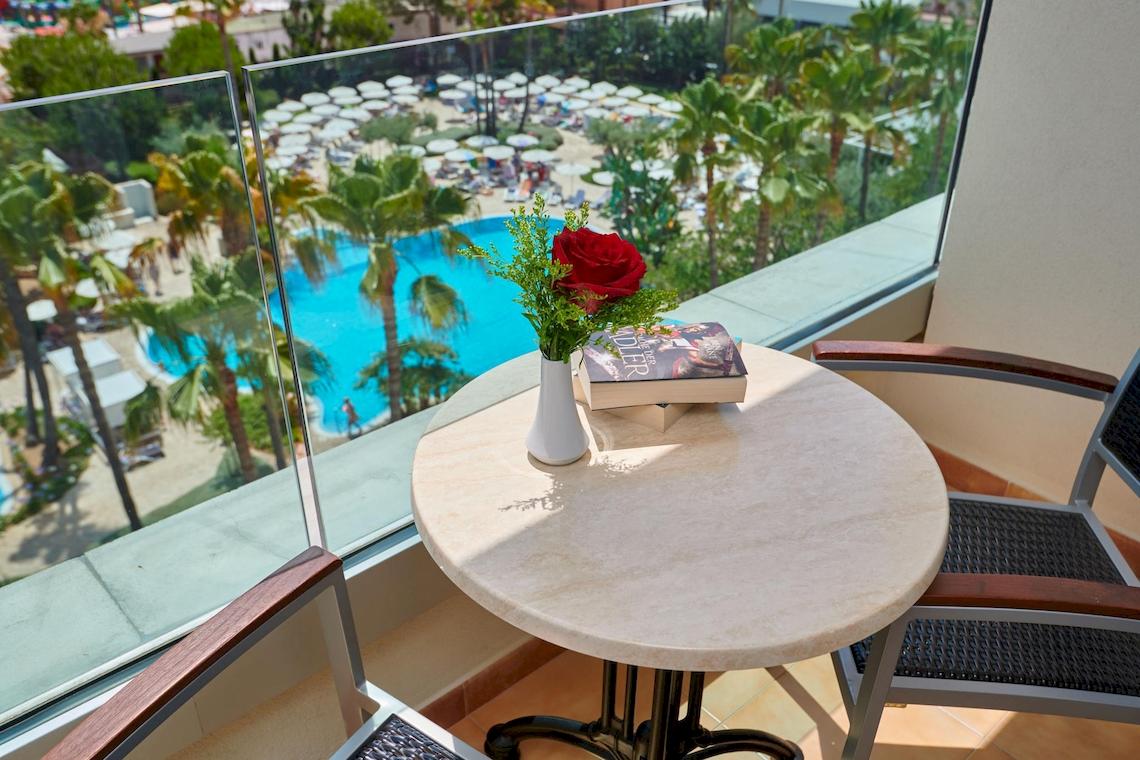 Blick vom Balkon des Hipotels Said mit Tisch, Buch und Rose auf der Hotelterrasse mit Pool im Hintergrund.