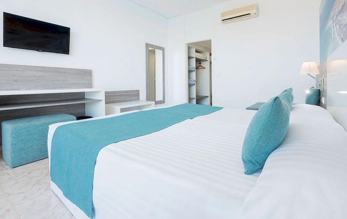 Schlafzimmer im Azuline Hotel Mar Amantis mit weißem Bett, blauen Kissen und Decken, Fernseher an der Wand, Schrank und Lampe.