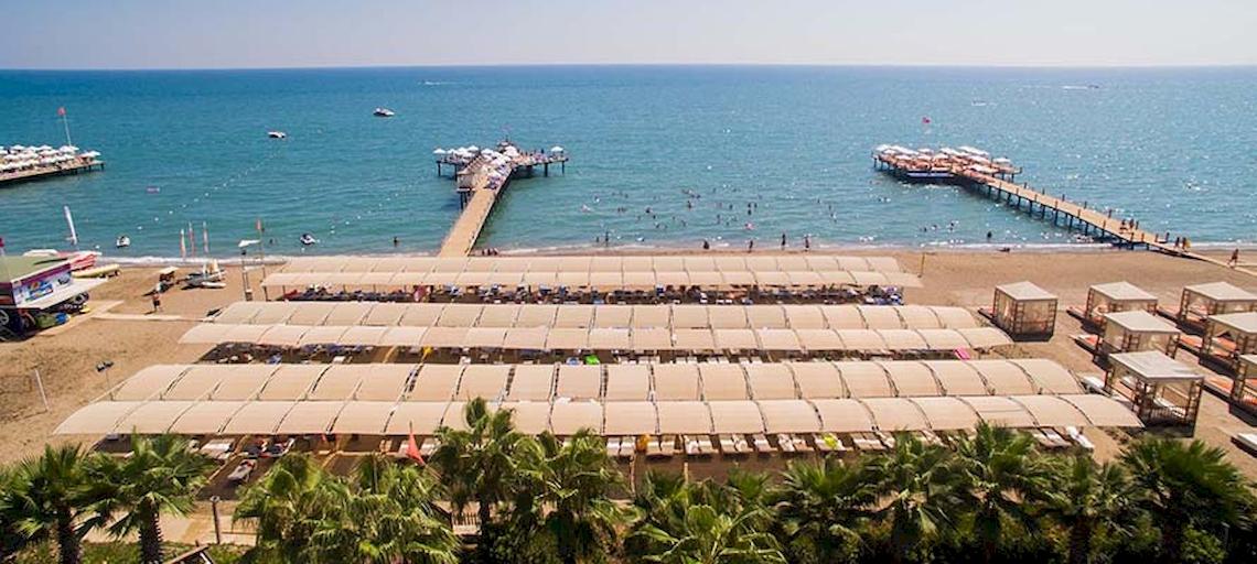Luftaufnahme des Saturn Palace Resorts Strands mit mehreren Sonnenschirmen und Liegen.